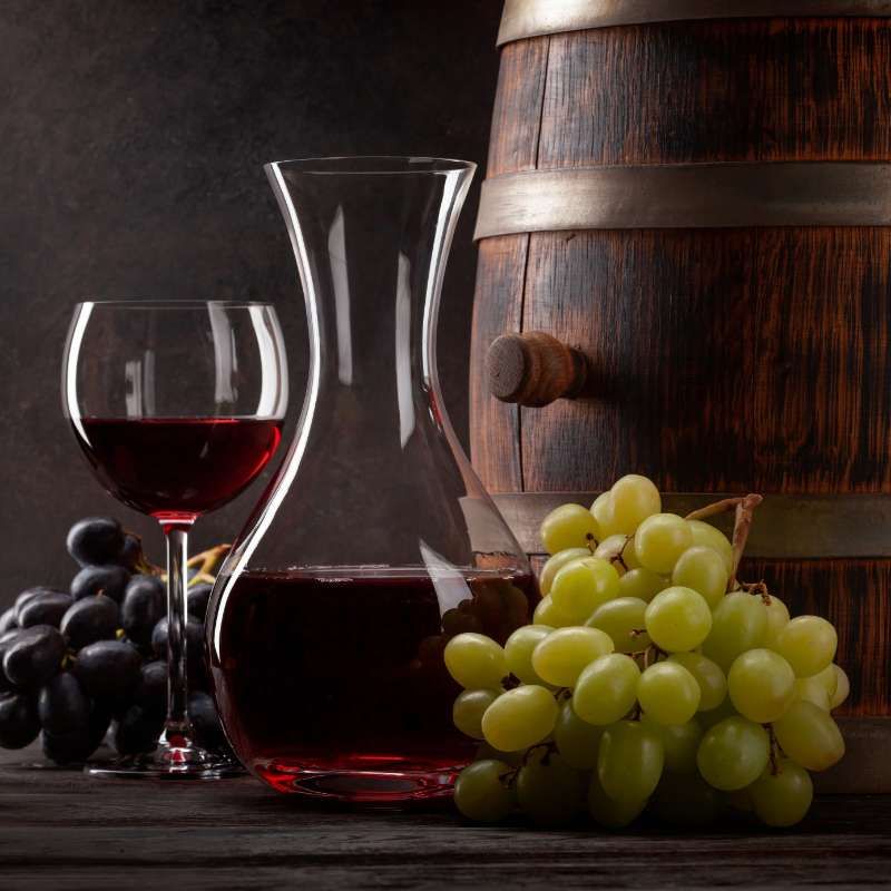Jarra y copa llenas de vino junto a uvas y un barril